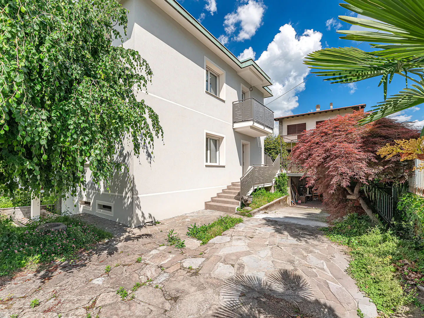 Two-story light gray house with a balcony, stone walkway, and green trees against a blue sky.