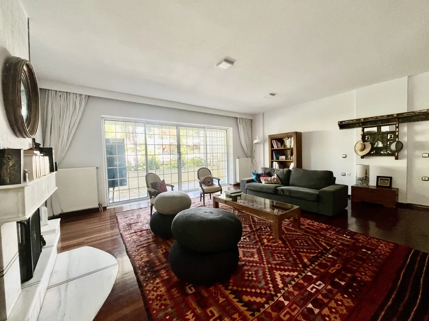 Bright living room with a red patterned rug, gray sofa, and a white fireplace. Large windows let in natural light.