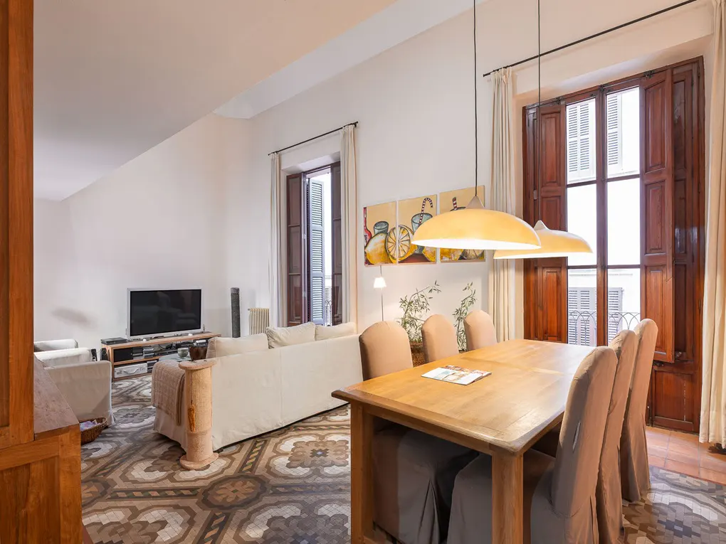 Bright living room with a wooden dining table, beige chairs, and a white sofa. Large windows with brown shutters and patterned floor tiles.