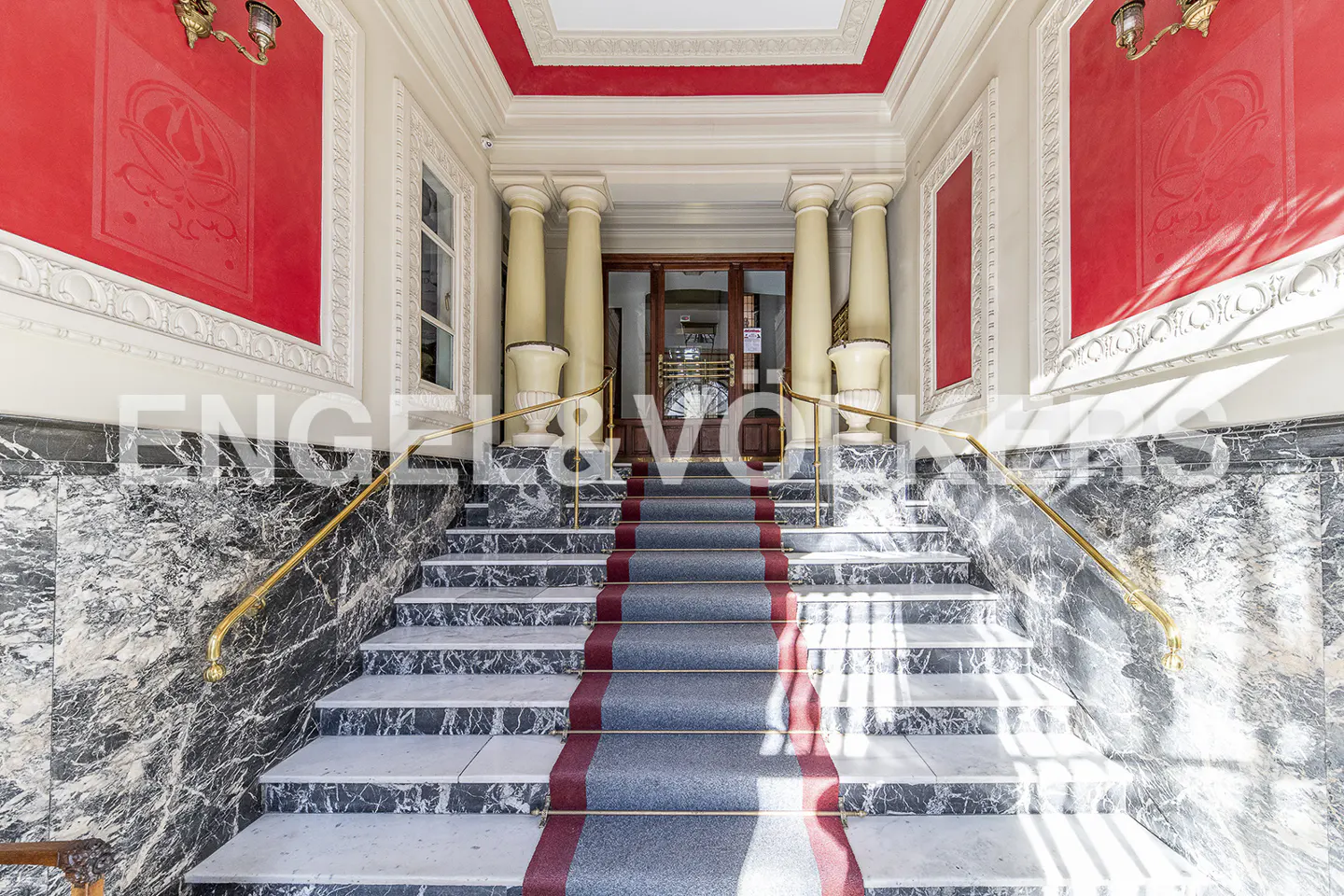 Grand entrance with marble stairs, gold railings, and a red-trimmed carpet leading to a wooden door framed by columns.
