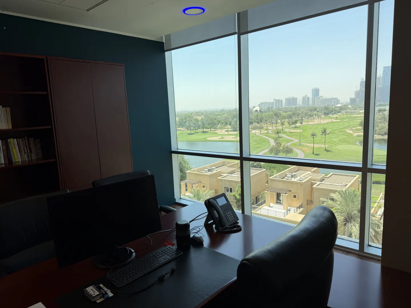 Office with a desk, computer, and phone. Large windows overlook a golf course, lake, and city skyline. The walls are blue, and there is a brown bookcase.