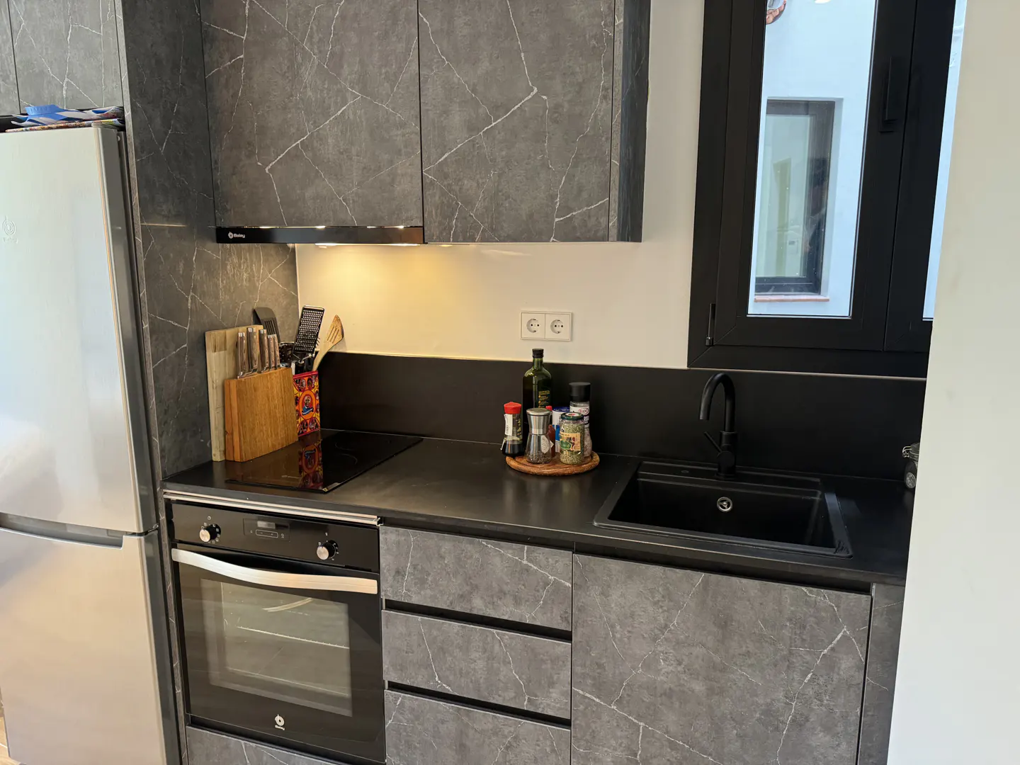 A modern kitchen with gray marble-patterned cabinets, black countertops, and a stainless steel refrigerator.