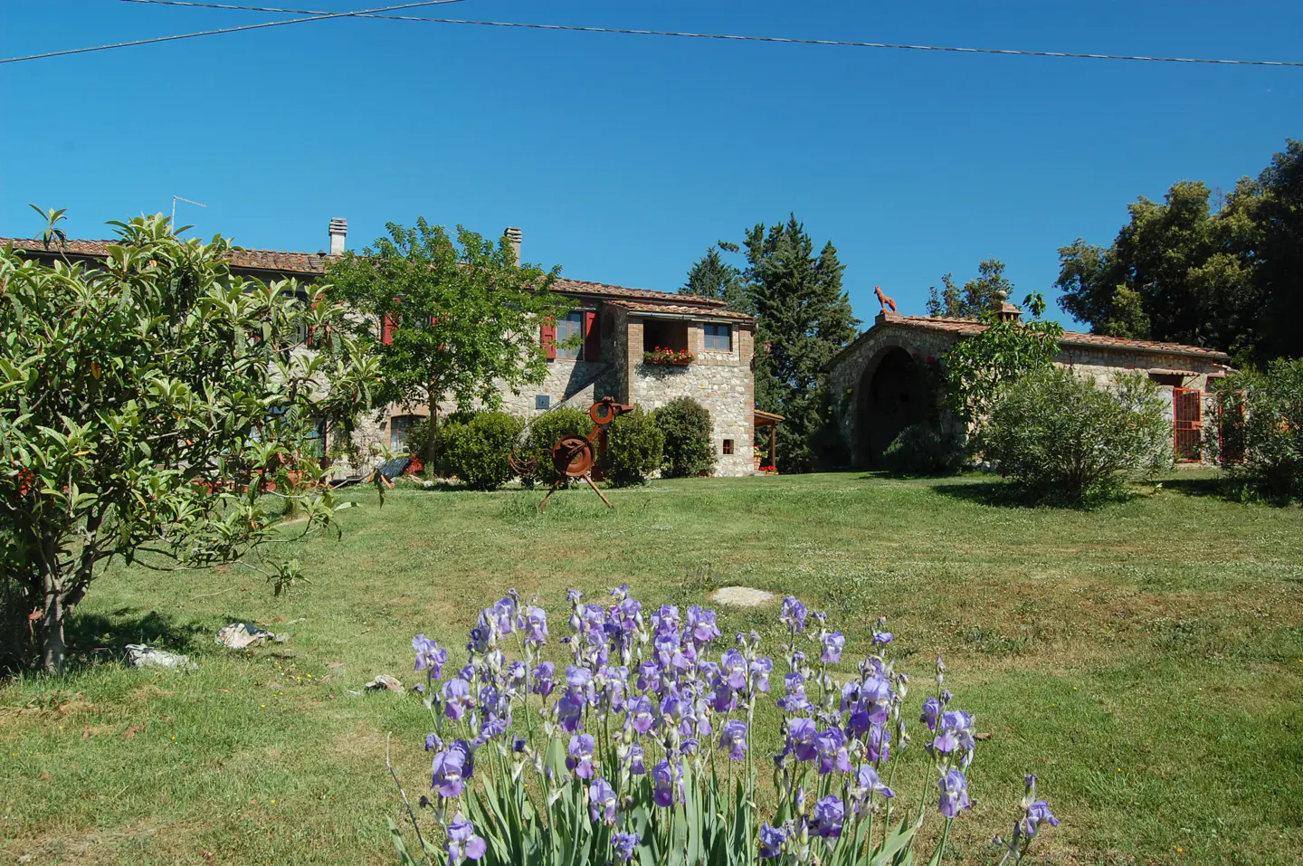 Stone house with red shutters and a red-roofed outbuilding, viewed across a lawn with purple irises under a clear blue sky.