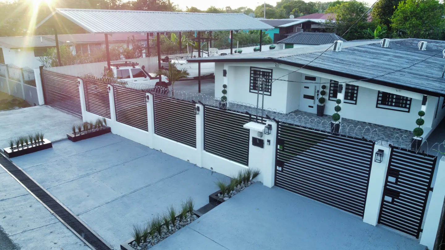 A white house with a black metal fence and gate, topped with barbed wire, sits on a concrete driveway.