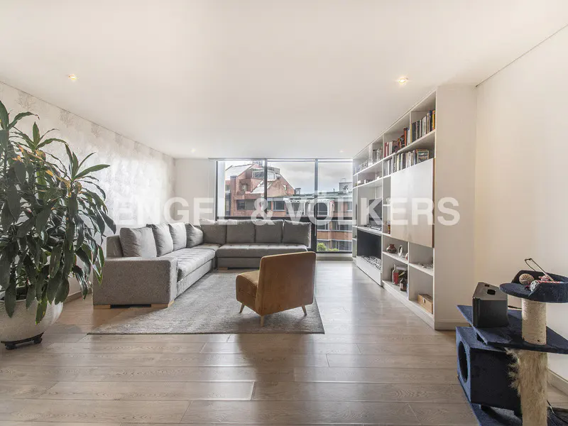 Bright living room with gray sectional sofa, brown chair, wood floors, and a large window with a city view. A tall bookshelf is on the right.
