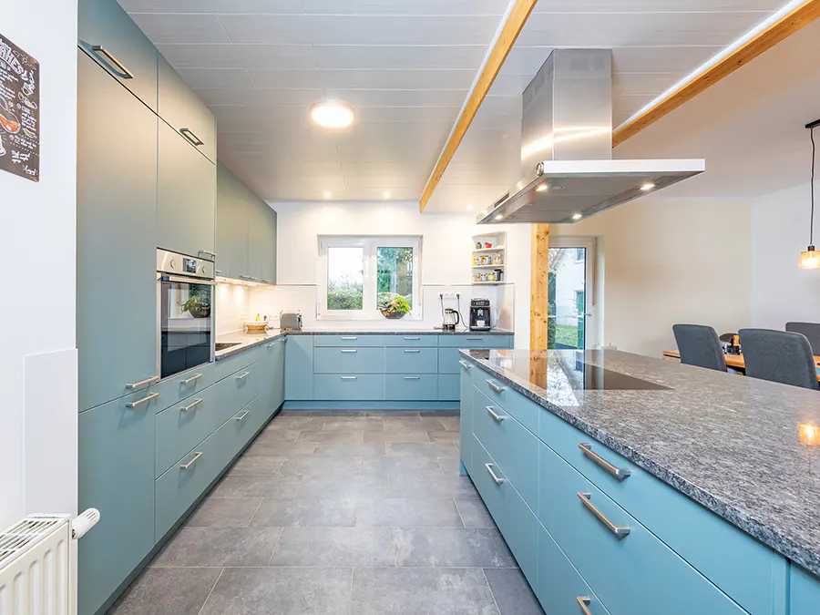 A modern kitchen with light blue cabinets, gray countertops, and gray tile flooring. A stainless steel range hood hangs above the island.