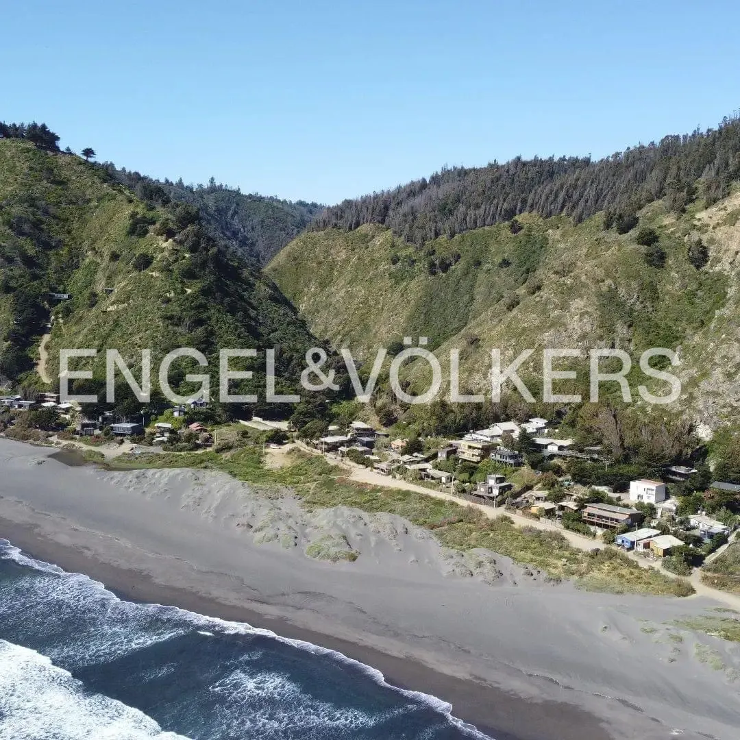 Aerial view of a coastal community nestled between a dark sand beach and steep, green, tree-covered hills.