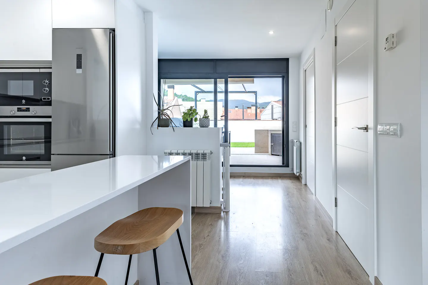 Modern white kitchen with stainless steel appliances, wooden bar stools, and a sliding glass door leading to a terrace.