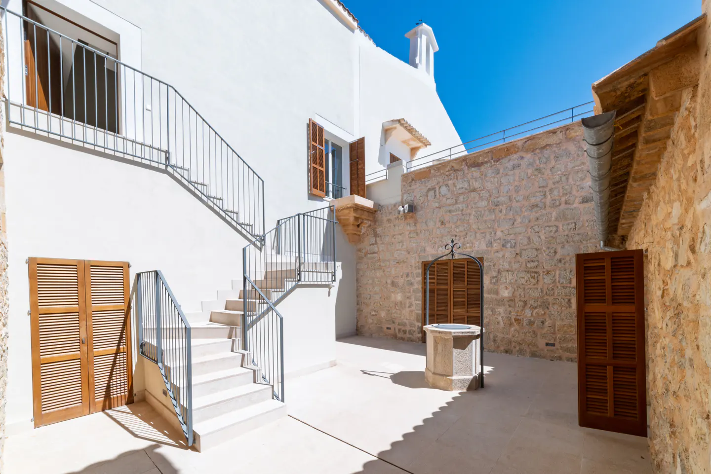 Exterior courtyard with white walls, stone accents, and a well. Stairs lead to an upper level. Shutters are closed on windows.