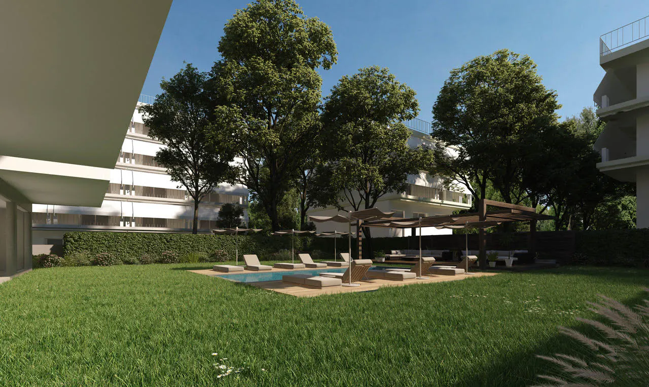 A swimming pool with lounge chairs on a wooden deck, surrounded by a green lawn and trees, set between modern buildings.