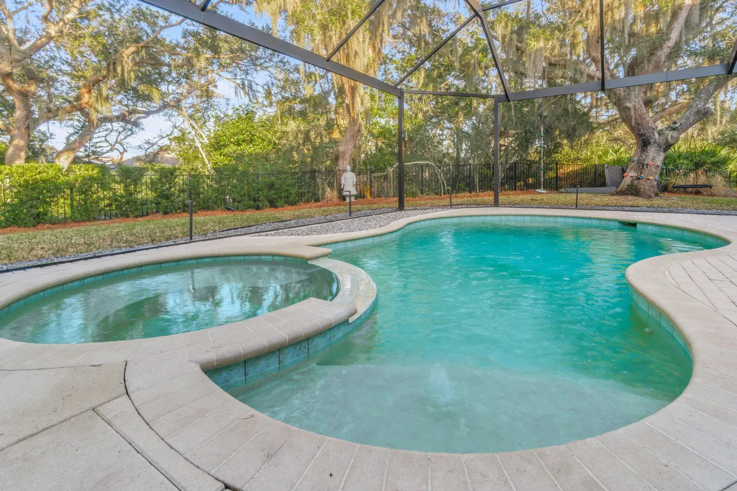 A kidney-shaped pool with a hot tub is surrounded by a screened enclosure and lush greenery.
