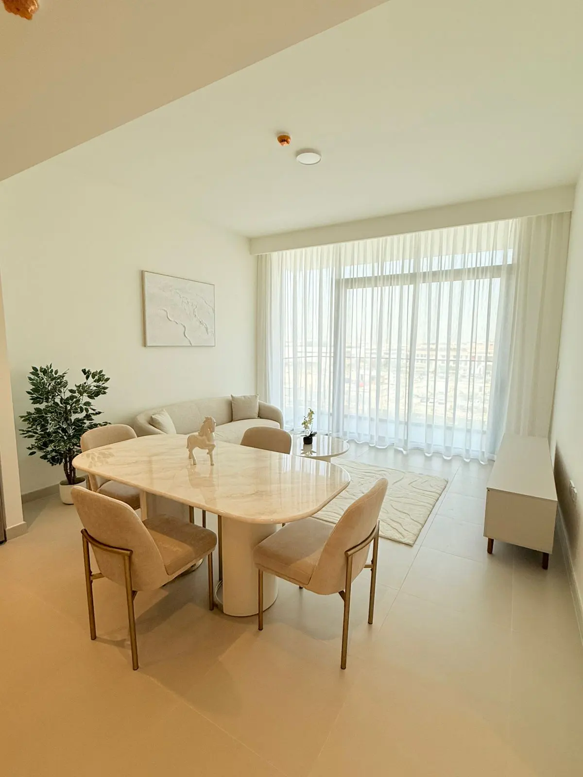 Modern dining area with a marble table, beige chairs, a potted plant, and floor-to-ceiling curtains in a bright room.