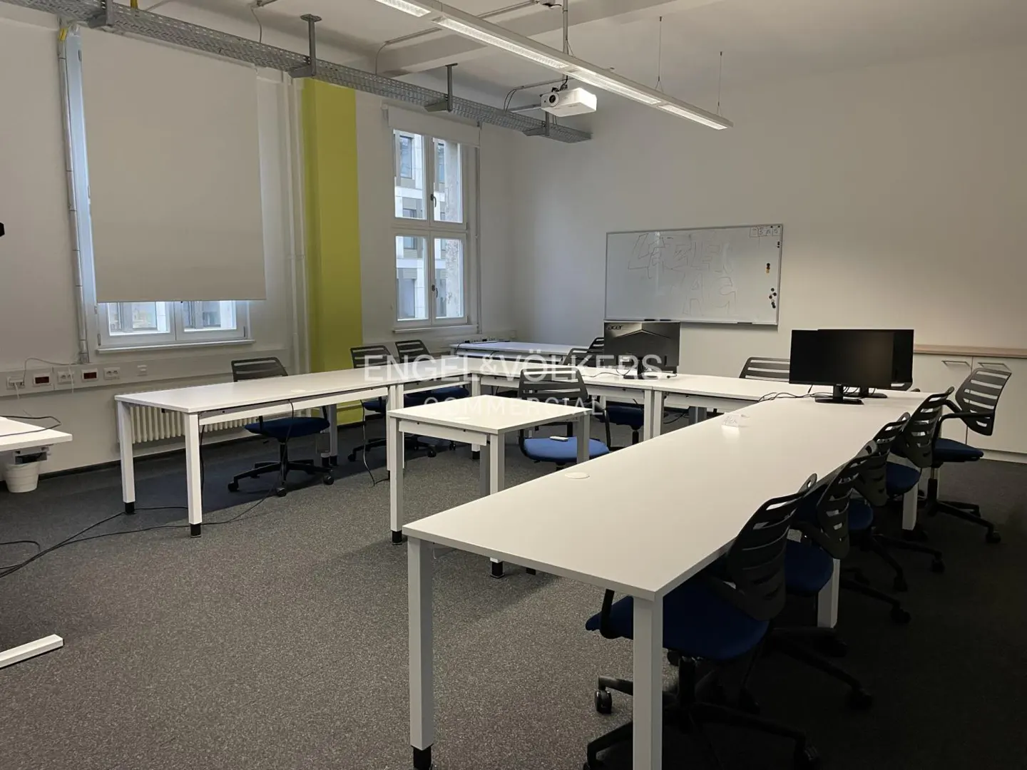 Bright classroom with white tables, blue chairs, and a whiteboard. A projector hangs from the ceiling. Natural light streams through the windows.