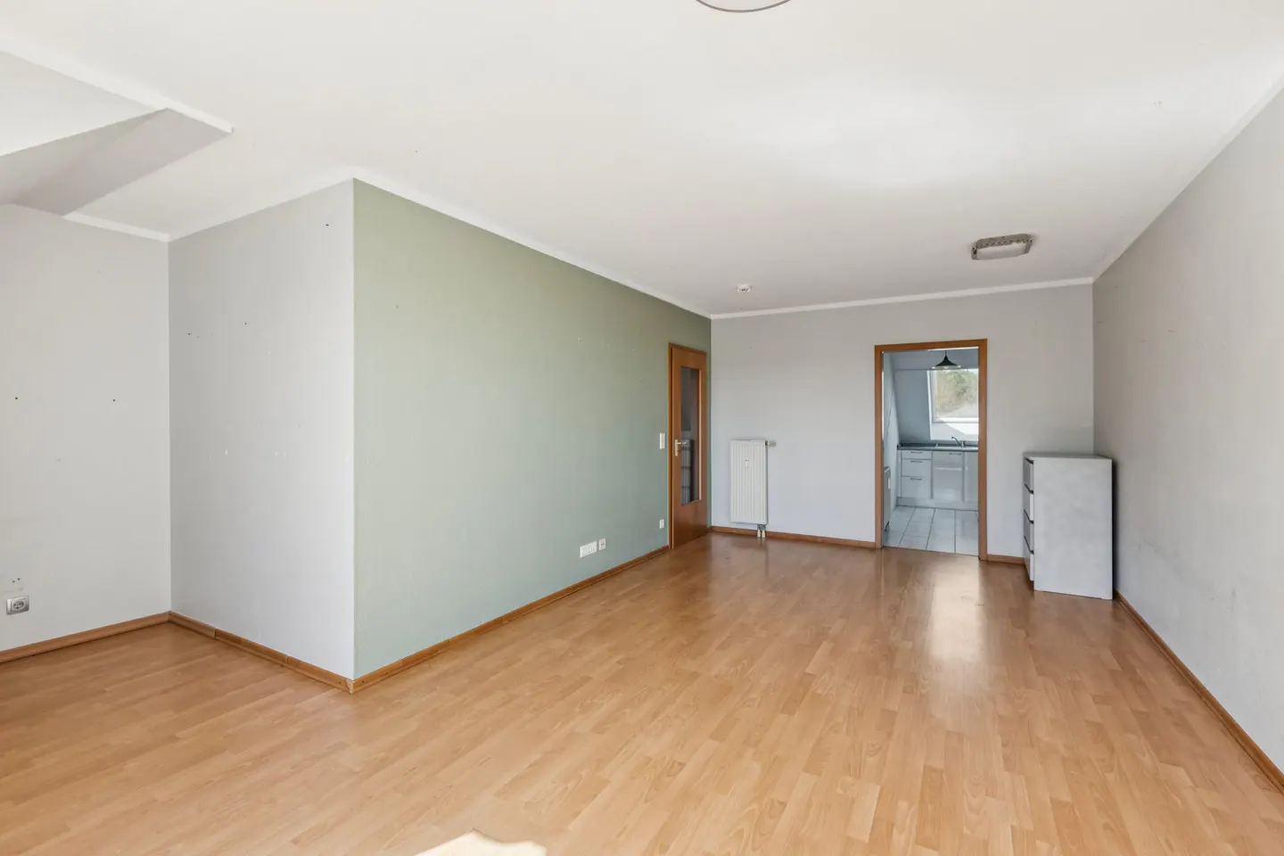 A bright, empty room with light wood floors, gray and green walls, and a doorway to a kitchen.