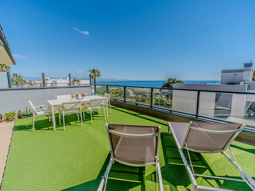 Rooftop patio with artificial grass, white table and chairs, and two lounge chairs overlooking the ocean on a sunny day.
