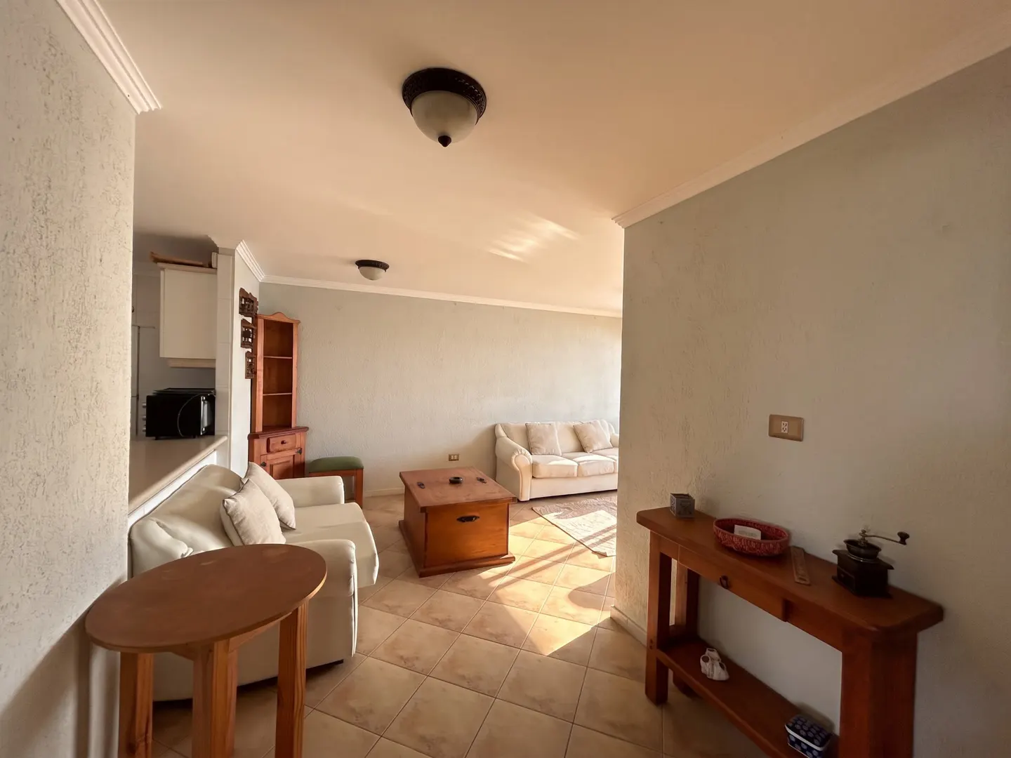 A bright living room with tile floors, light blue walls, and white sofas. A wooden coffee table and console table are also visible.