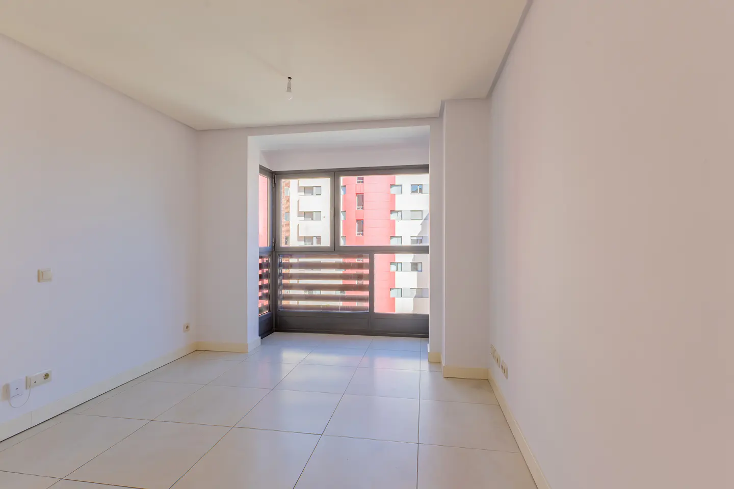 Empty room with white walls and tile floor. A large window shows a red and white building.