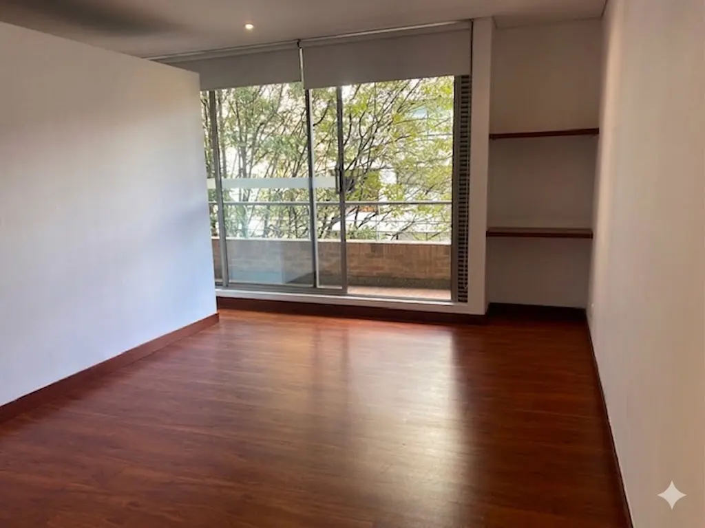 Empty room with hardwood floors, white walls, and a large window with a view of trees and a balcony. Built-in shelves are on the right wall.
