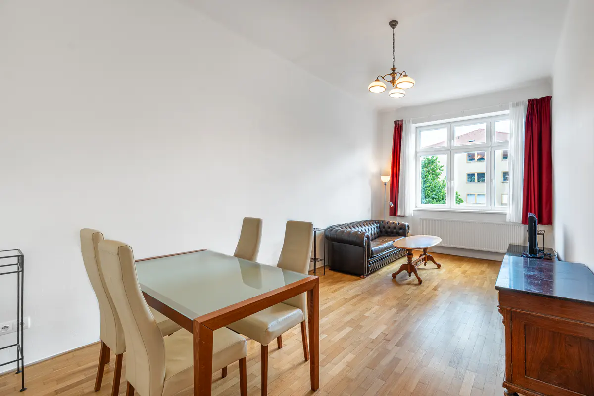 Bright living room with a glass dining table, four beige chairs, a black leather sofa, and red curtains by the window.