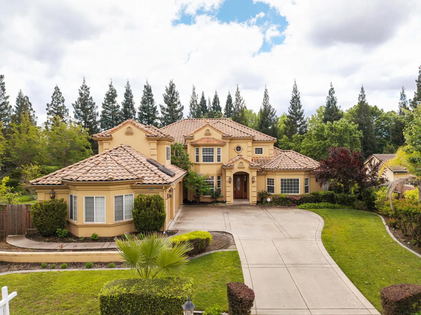 Beige two-story house with a tile roof, green lawn, and a long driveway on a cloudy day.