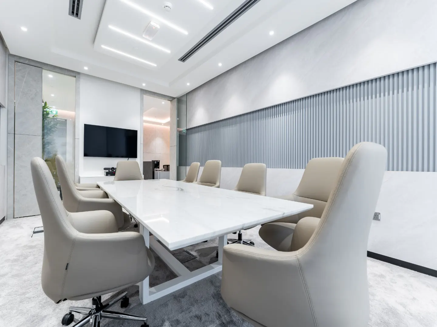 Modern conference room with a white marble table, beige leather chairs, and a wall-mounted TV in a bright, minimalist office.