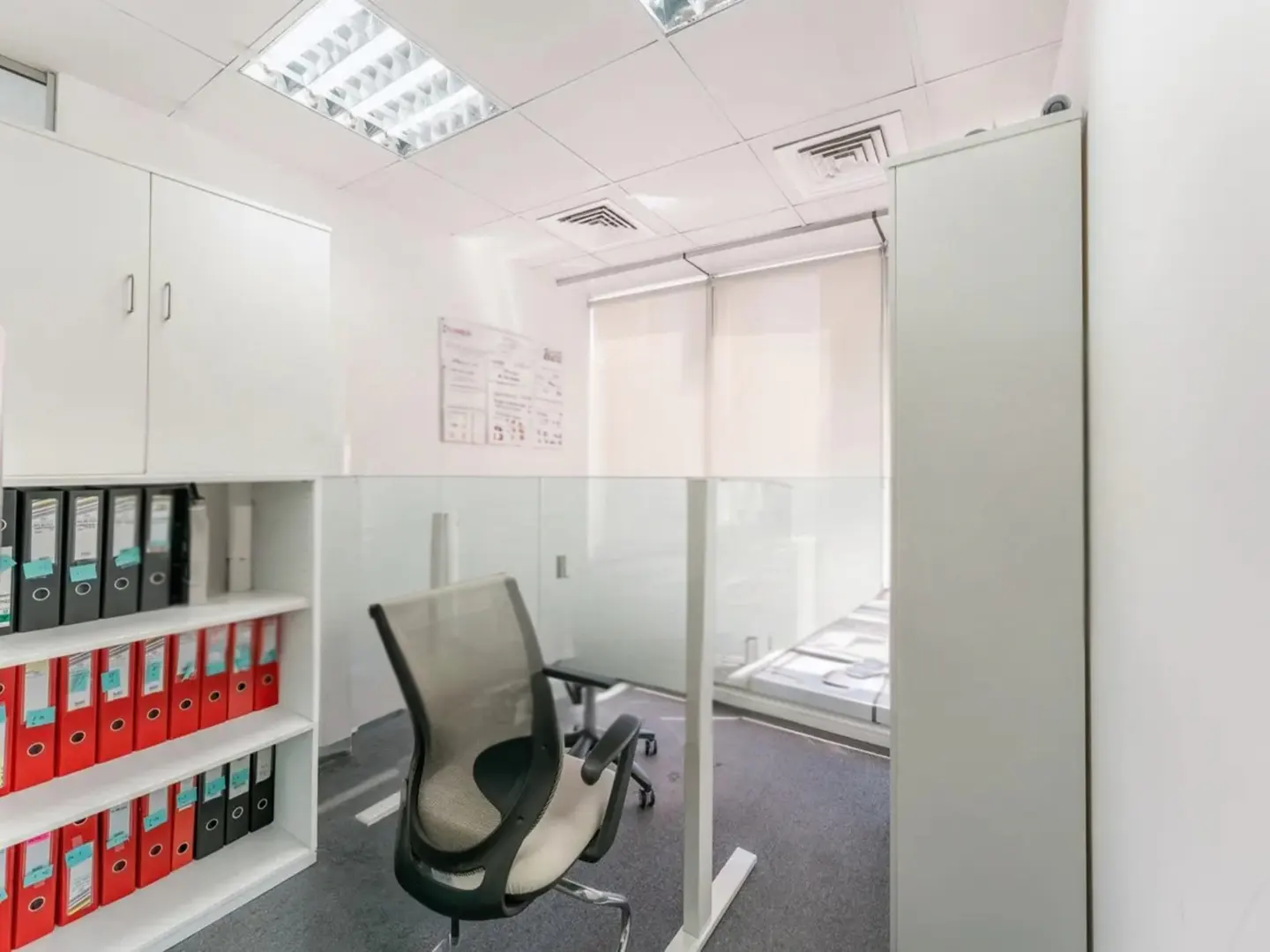 Office space with white walls, cabinets, and a glass-enclosed cubicle with a gray office chair. Red and black binders are on shelves.