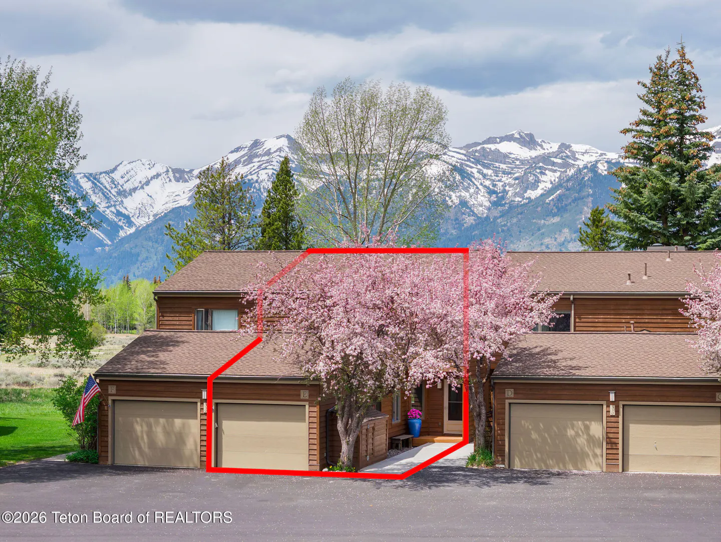 Two-story brown townhouse with garage doors, framed in red, with pink flowering trees and snow-capped mountains in the background.