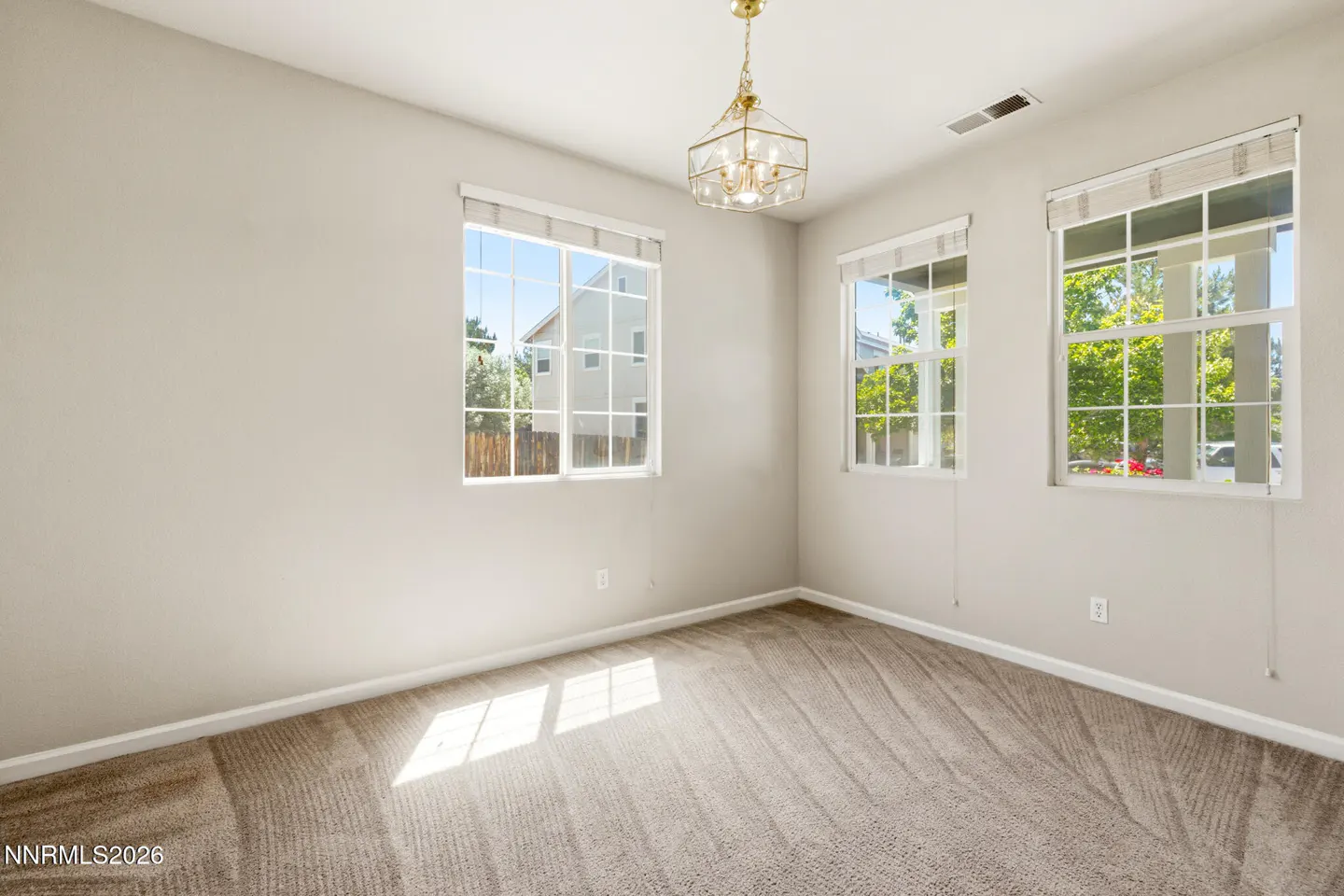 Empty room with beige walls, tan carpet, three windows with blinds, and a gold geometric pendant light fixture.