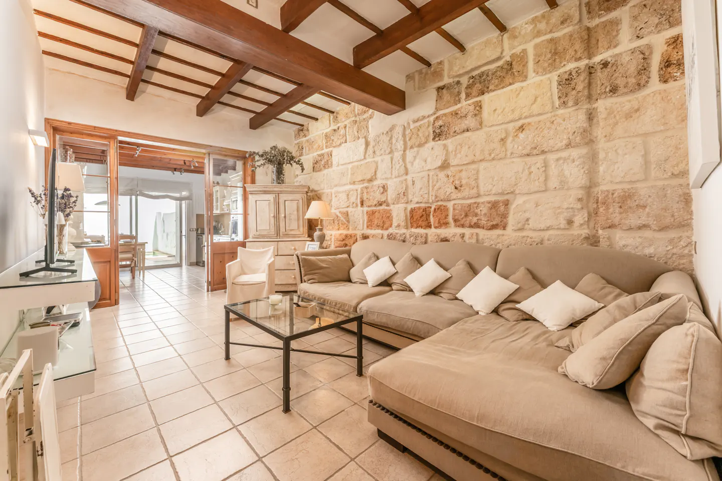 Living room with beige sectional sofa, stone wall, and wooden ceiling beams. Glass table and white armchair are also visible.