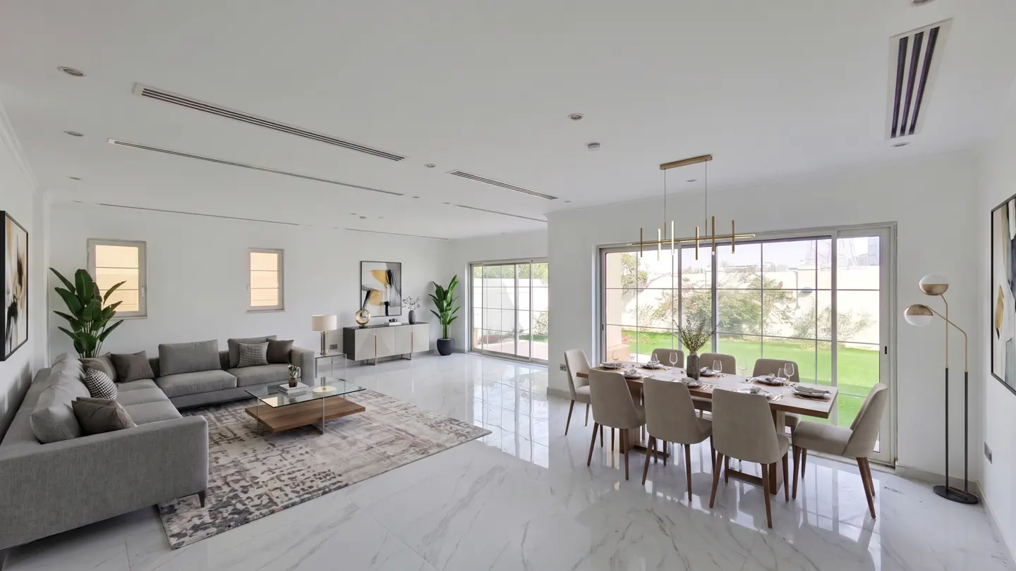 Bright, open-plan living and dining area with white walls and marble floors. A gray sectional sofa faces a glass coffee table and a dining table set for eight sits near a large window.