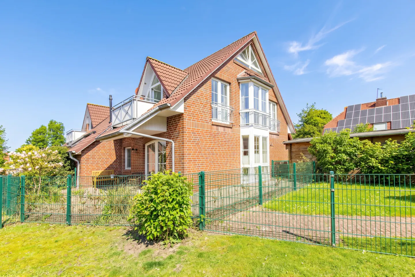 Two-story brick house with a red tile roof, white trim, and a green metal fence on a sunny day.