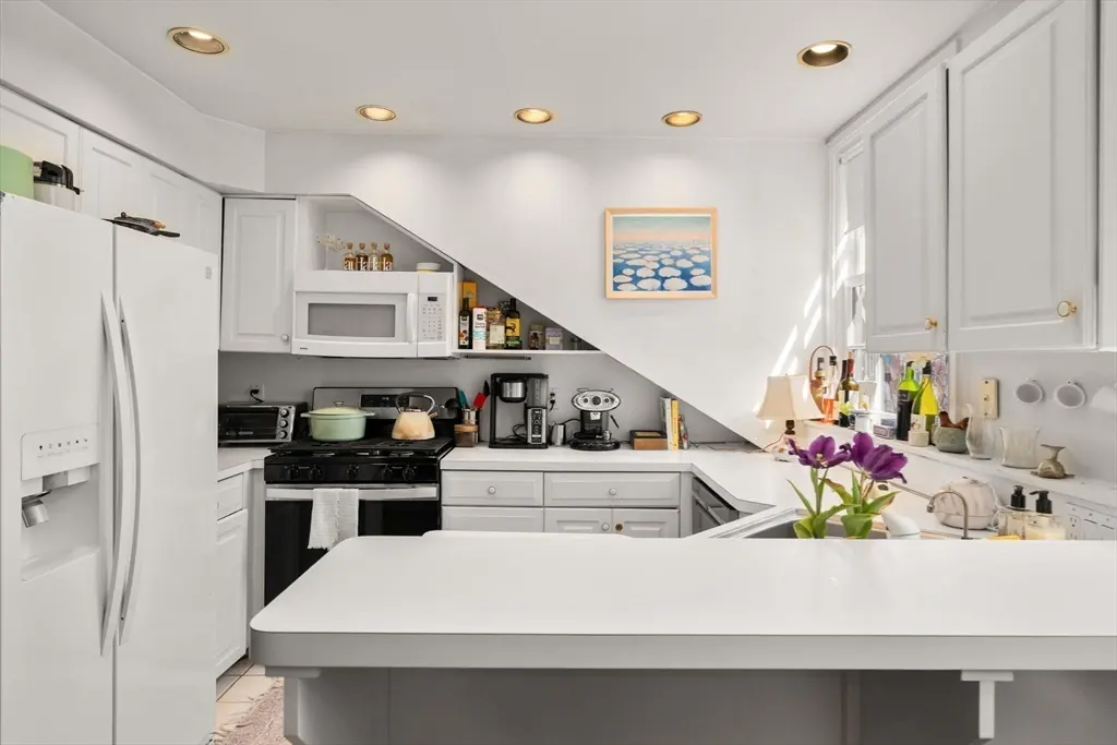 Bright kitchen with white cabinets, appliances, and countertops. A painting of clouds hangs on the wall. Purple tulips sit on the counter.