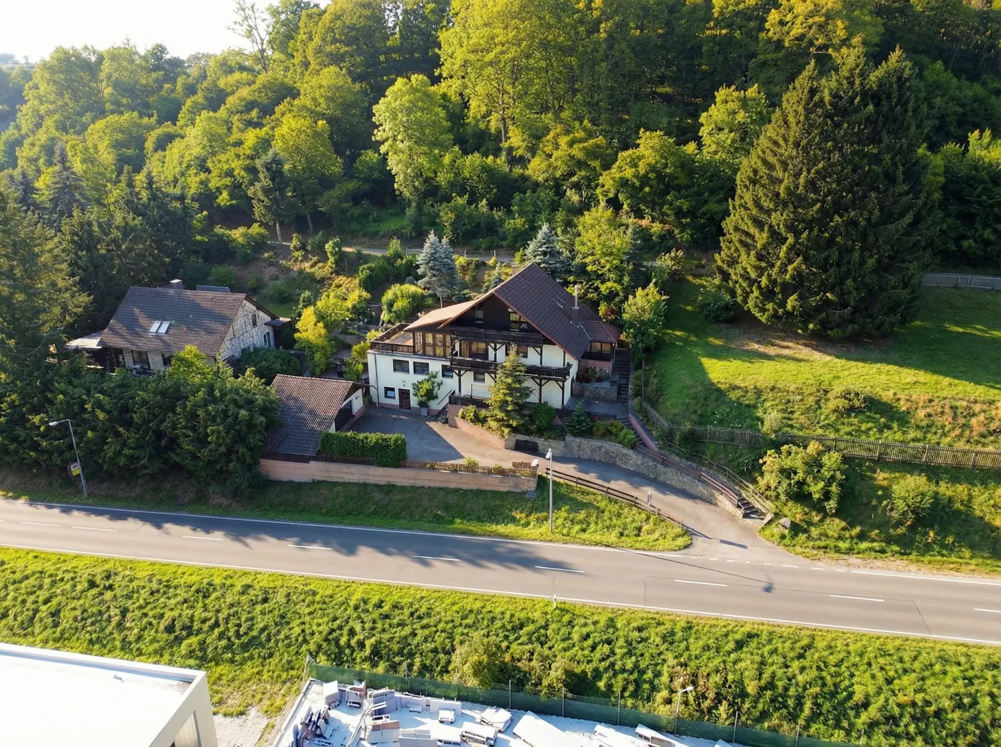 Aerial view of a white two-story house with a brown roof, nestled on a green hillside with lush trees. A road runs in front of the property.