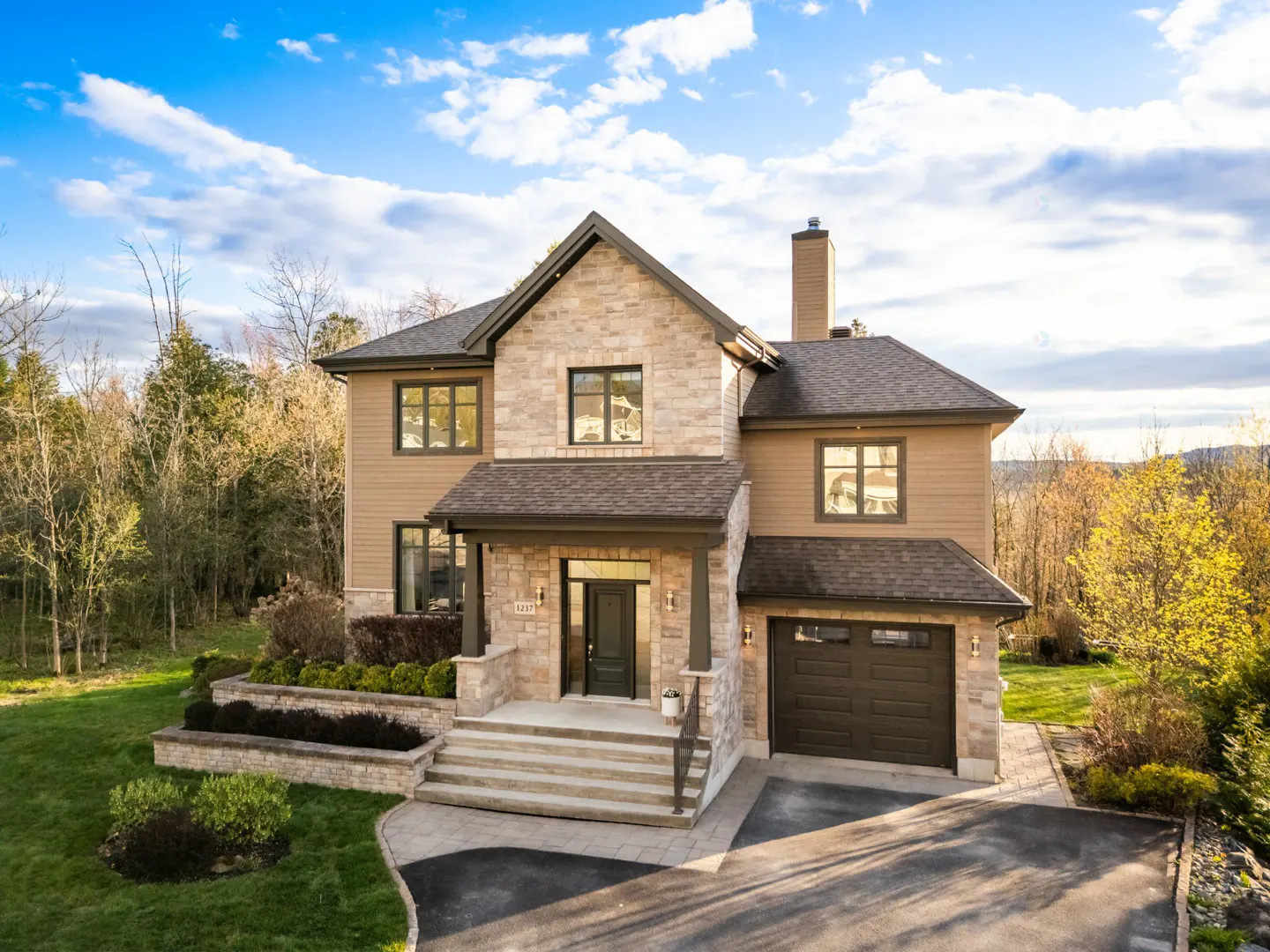 Two-story house with stone and tan siding, a dark roof, and a two-car garage. Landscaped front yard with stone retaining walls and green grass. Blue sky with clouds.