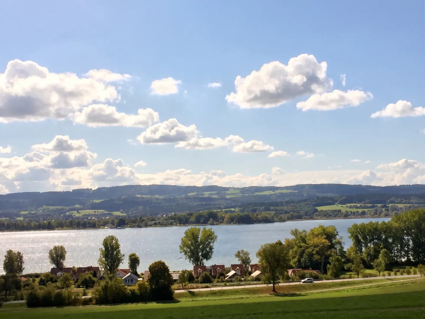 Scenic view of a lake with houses, trees, and a road in the foreground. Rolling hills and a blue sky with white clouds in the background.