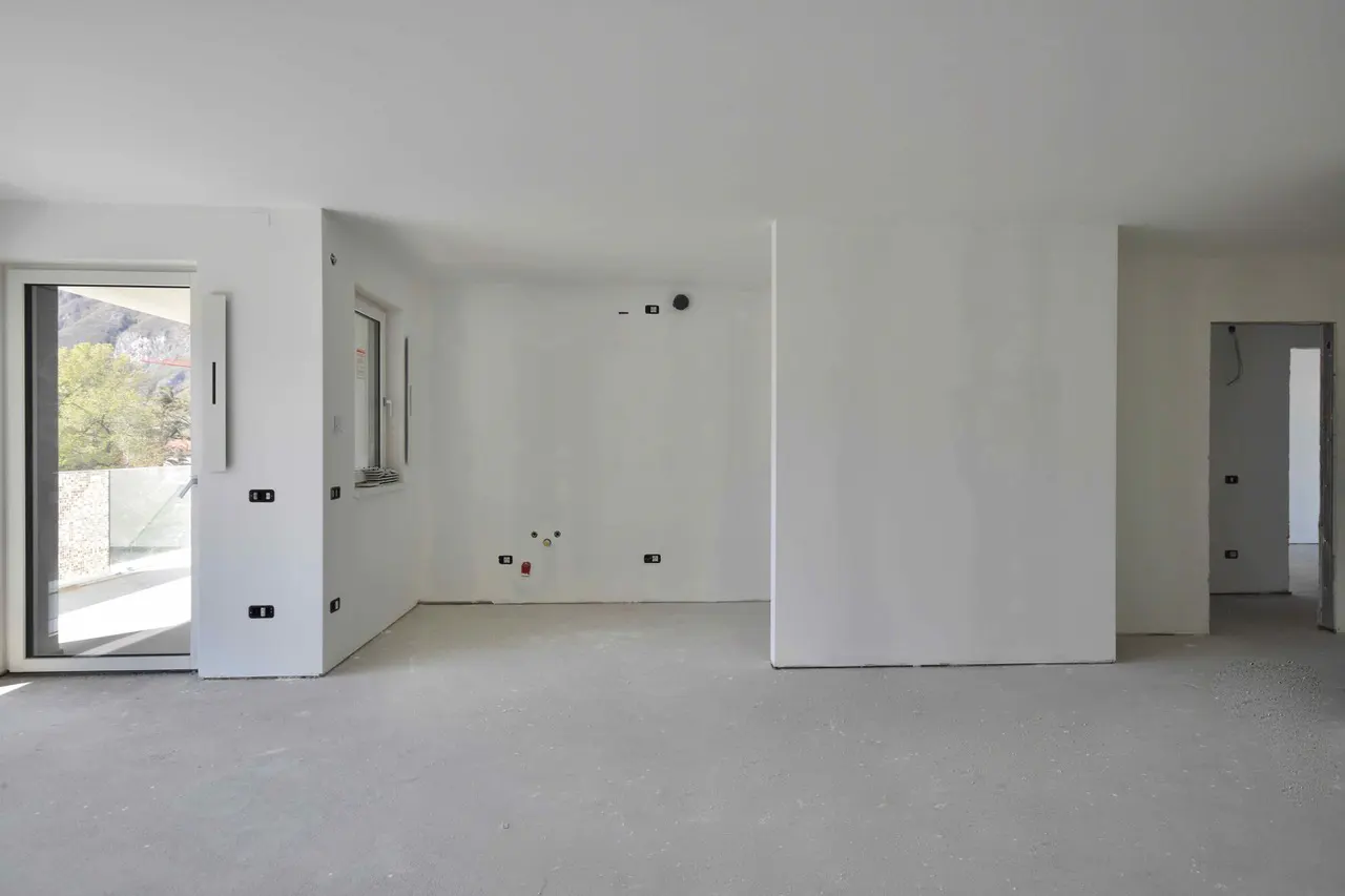 Unfinished room with white walls, grey floor, window, and sliding glass door to a balcony. Electrical outlets are visible.