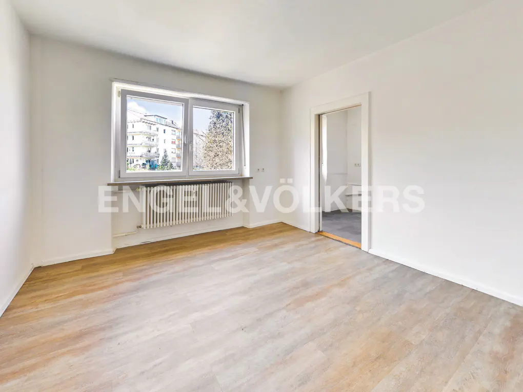 Bright, empty room with light wood floors, white walls, and a window overlooking a building and trees. A doorway leads to another room.