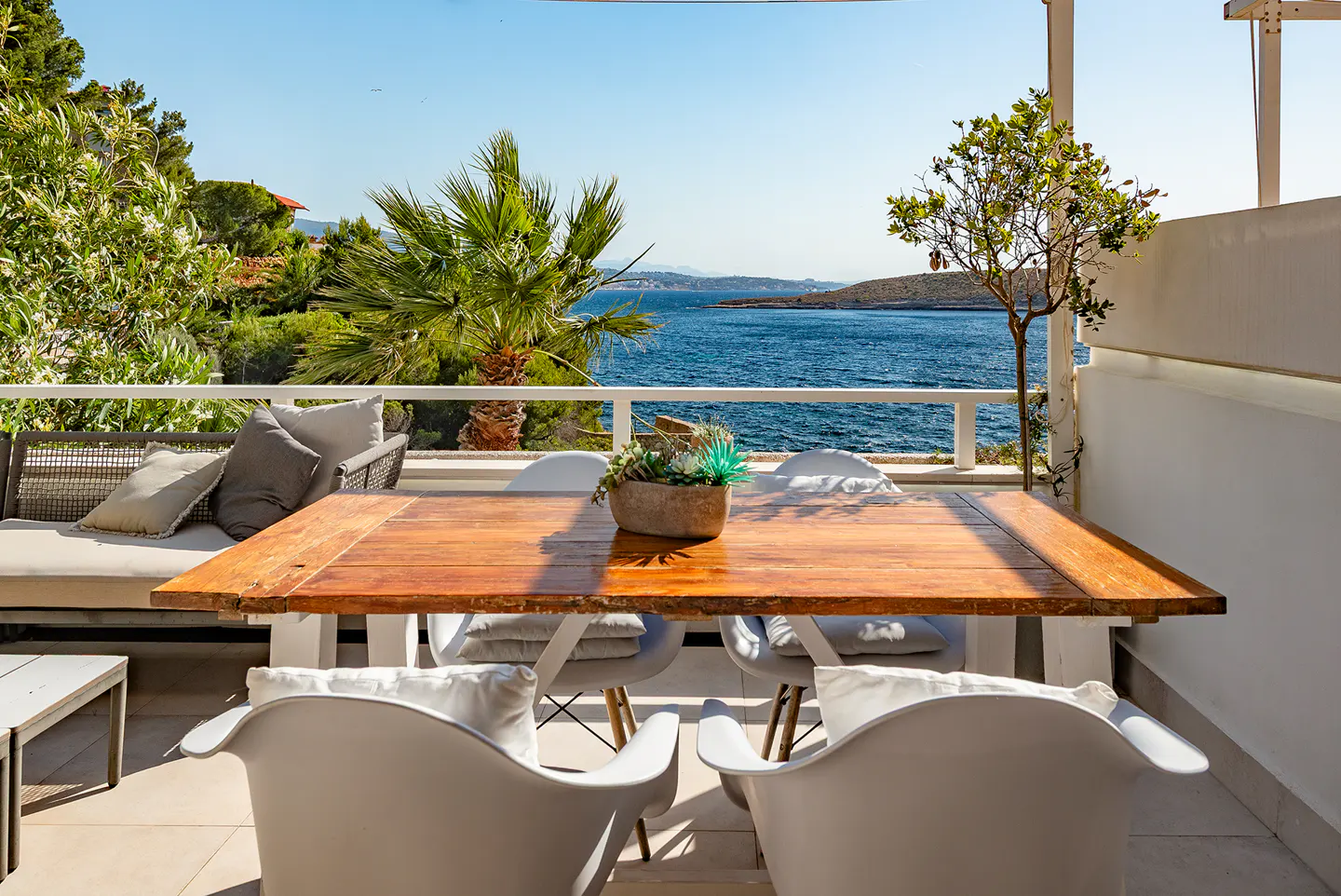 Sunny terrace with a wooden dining table, white chairs, and a sofa overlooking a blue sea and coastal landscape.