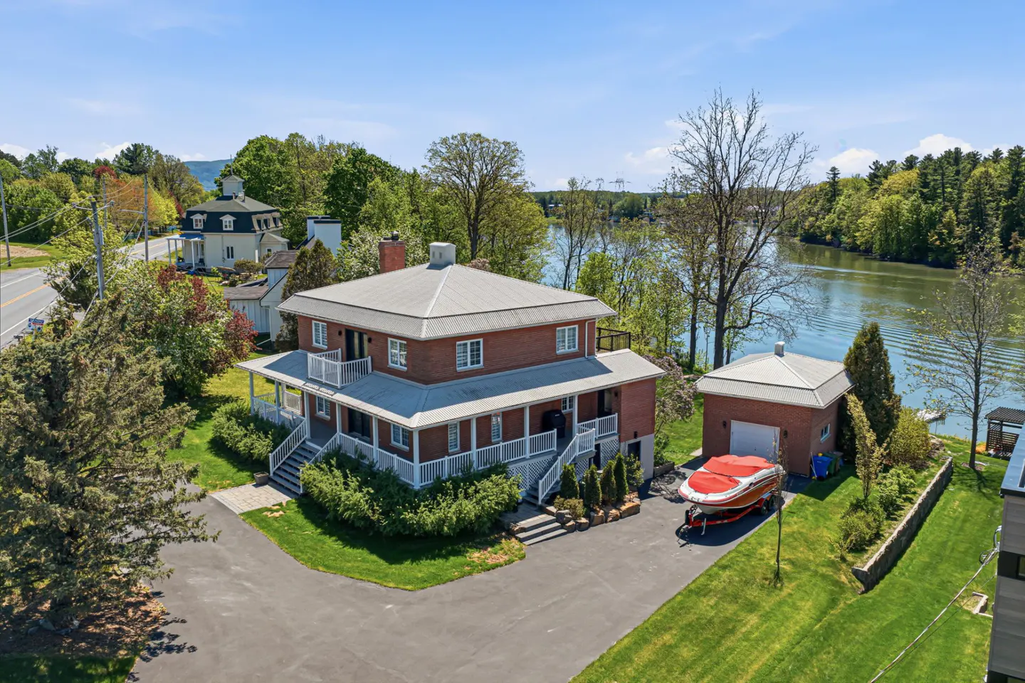 Aerial view of a two-story brick house with a gray roof, white trim, and a wraparound porch, near a lake and a boat on a trailer.