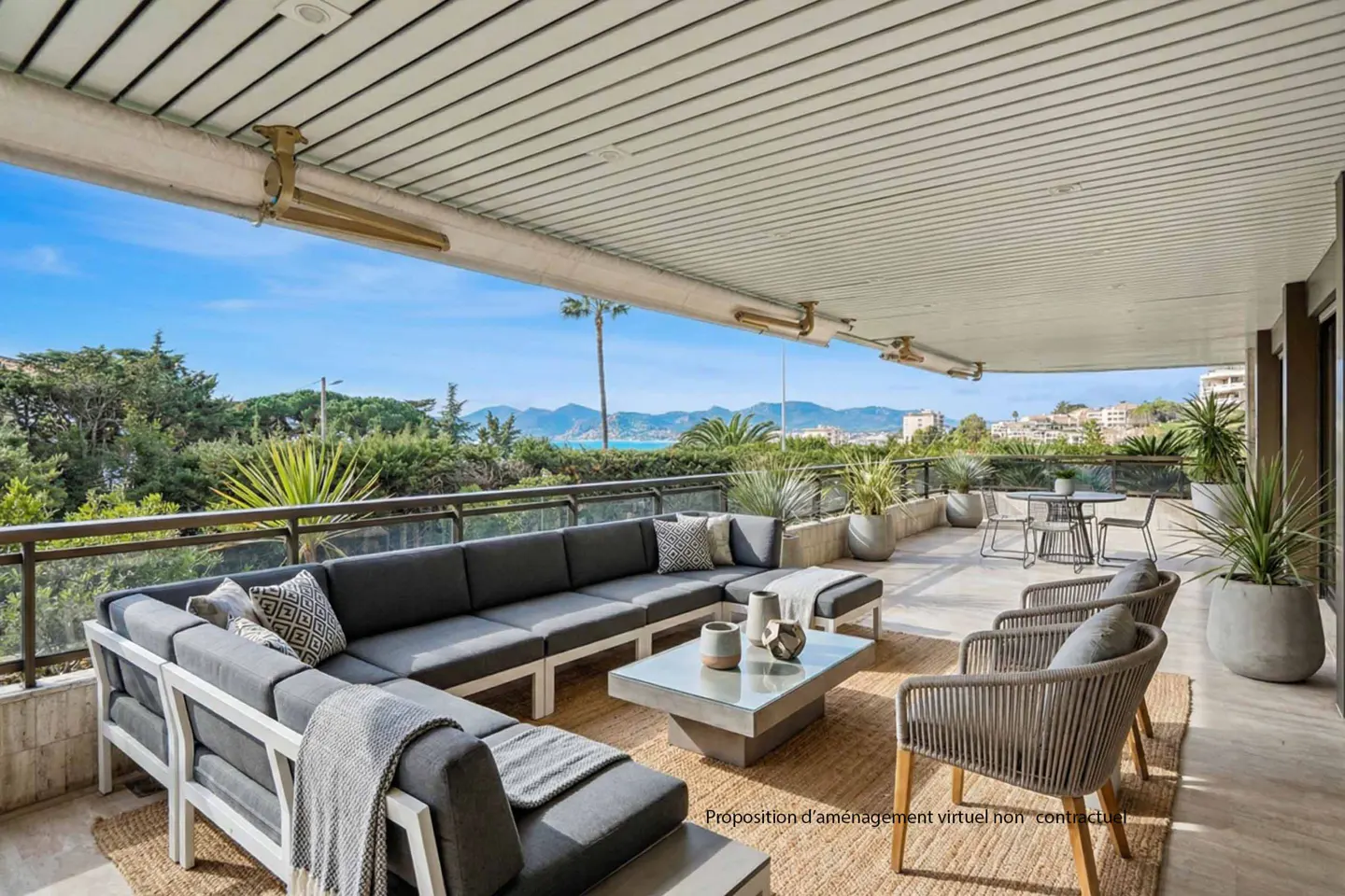 A covered patio with a gray sectional sofa, chairs, and a table overlooking a scenic view of trees, buildings, and the ocean.