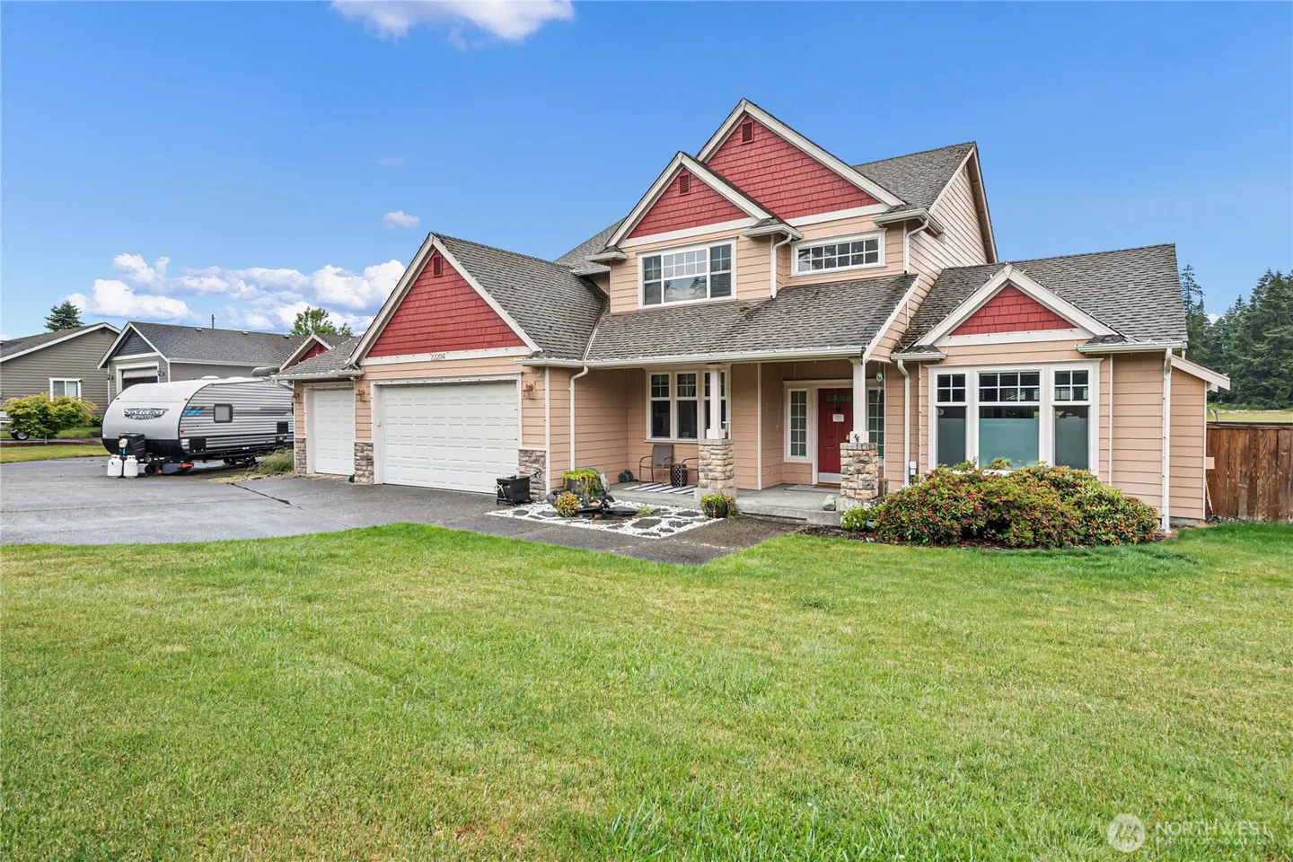 Two-story tan house with red accents, a three-car garage, and a travel trailer parked in the driveway on a sunny day.