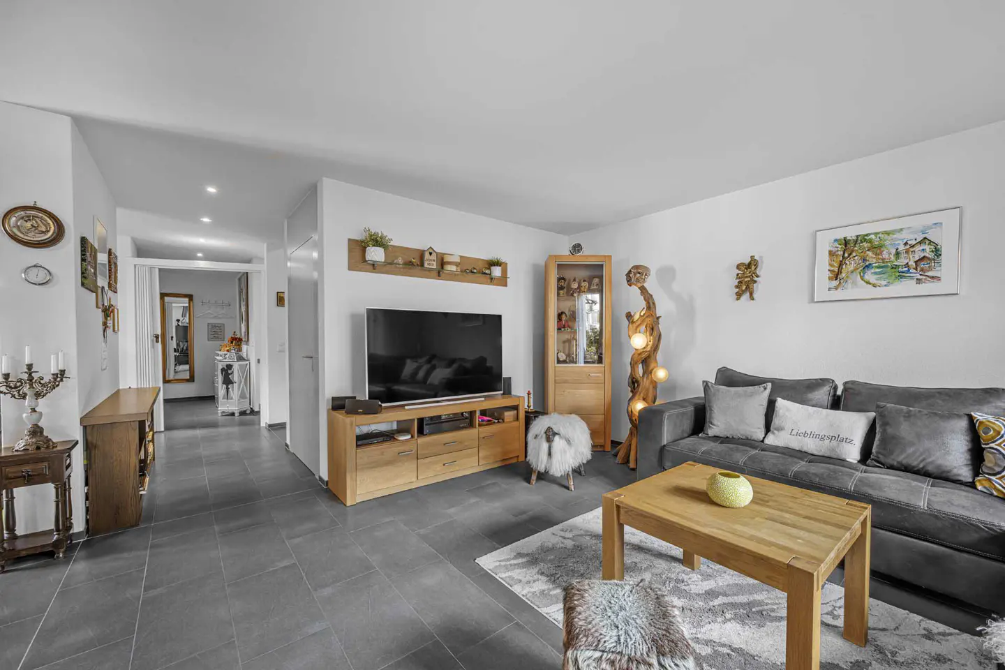 Living room with gray tile floor, gray sofa, wooden TV stand, and hallway leading to another room.