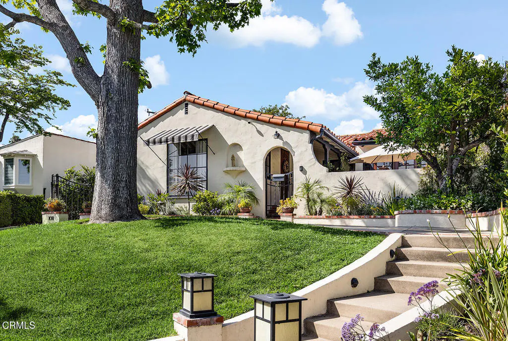 Exterior of a cream stucco house with a red tile roof, green lawn, and a large tree. Steps lead up to the house.