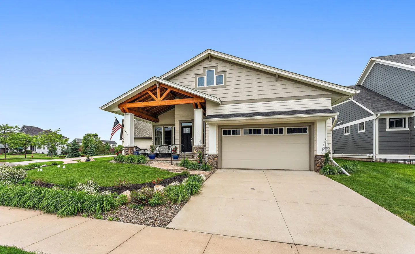 Beige single-family home with a front porch, American flag, and a two-car garage on a sunny day.