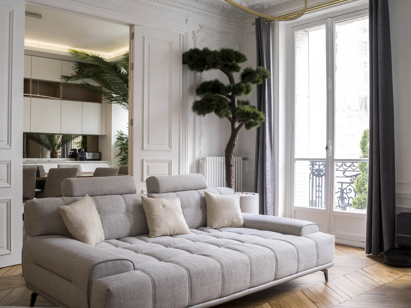 Modern living room with a light gray tufted sofa, a decorative bonsai tree, and a view into a kitchen with parquet floors.
