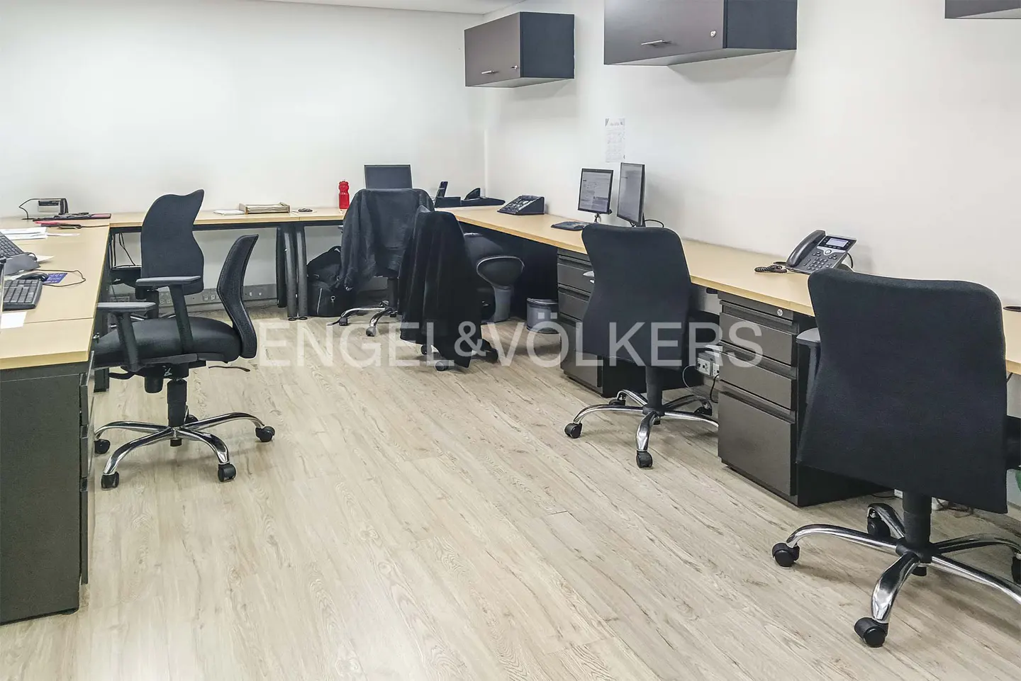 Office space with desks, black chairs, computers, and phones. Light wood floors and white walls. Cabinets are mounted on the wall.