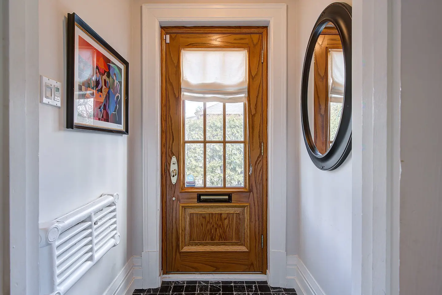 Entryway with a wood door, white walls, and black tile floor. A framed painting and a white radiator are on the left wall. An oval mirror hangs on the right wall.