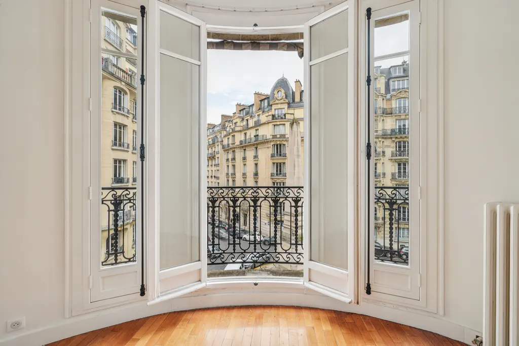 Interior view of a room with hardwood floors and white walls, featuring an open window with a wrought iron balcony overlooking a Parisian street.