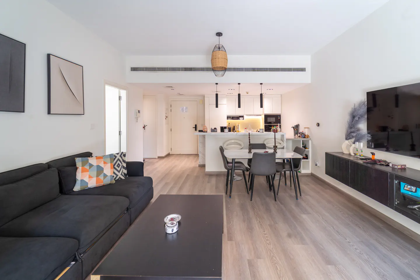 Open-concept living space with a black sofa, coffee table, dining set, and white kitchen. Artwork adorns the walls.