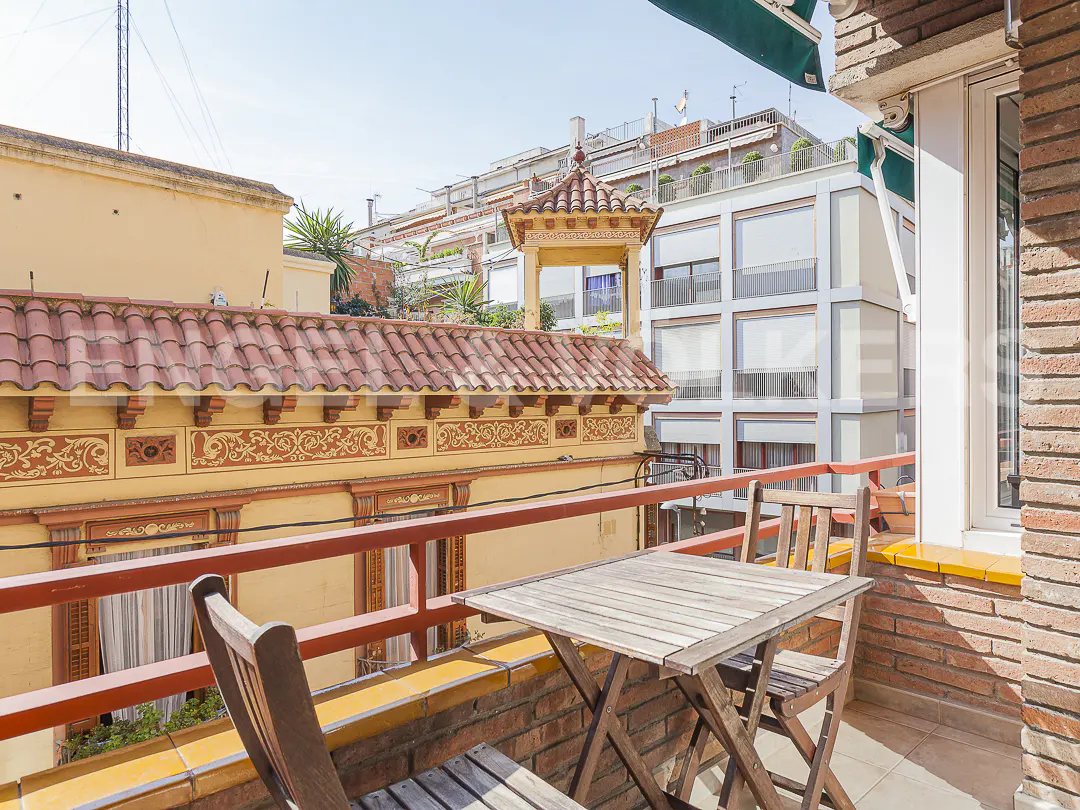 Balcony view with wooden table and chairs, red railing, and brick wall. Buildings with red tile roofs and white facades in the background.
